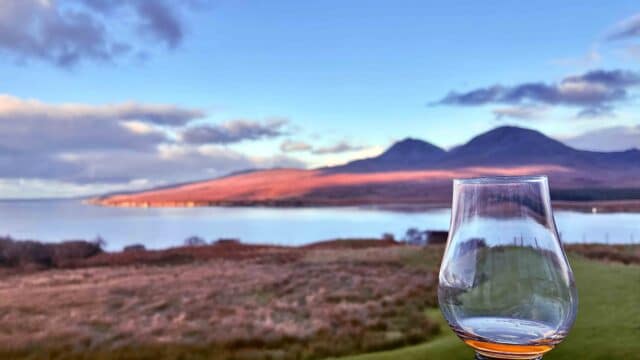 A picture of a whisky dram behind coastal scenery at Ardnahoe Distillery on Islay